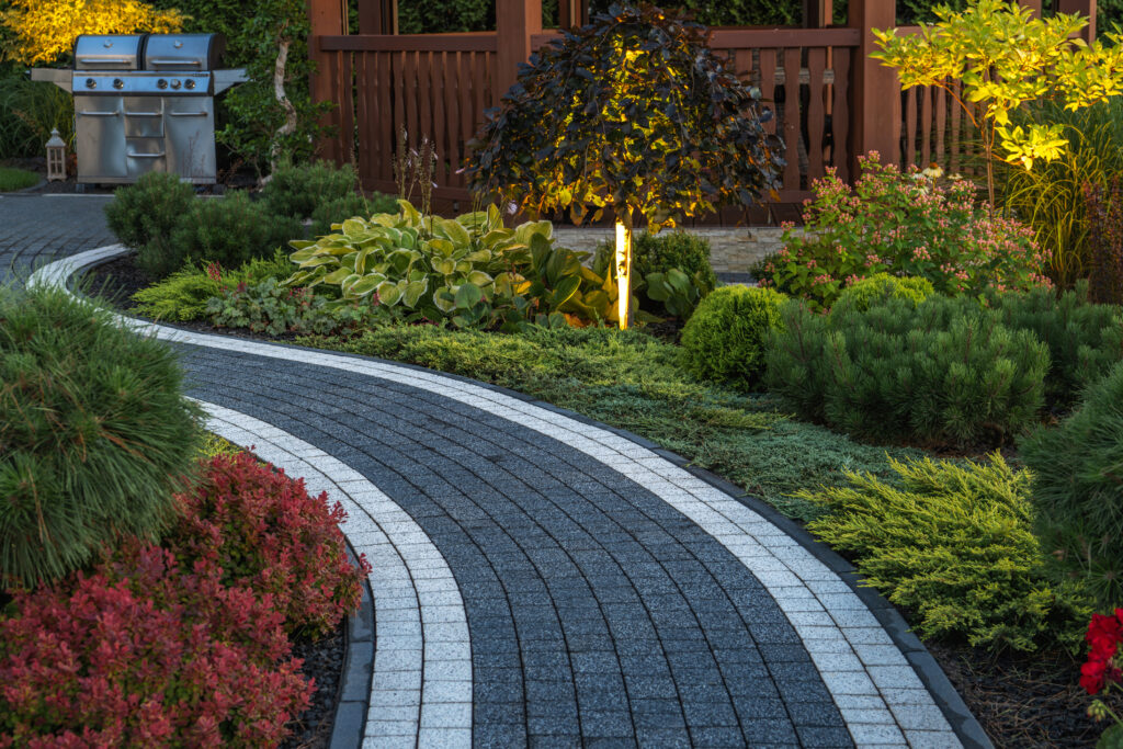 A curved concrete brick garden pathway surrounded by greenery in a backyard with a grill and gazebo in the background.
