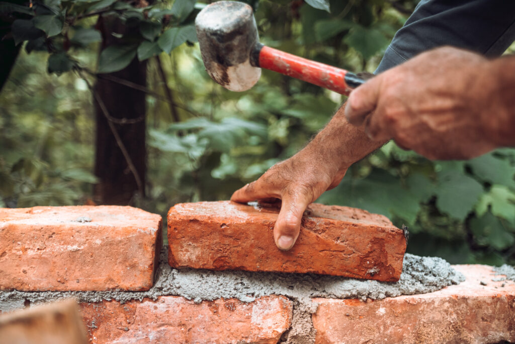 Closeup of a worker with a hammer placing a red brick on a wall.